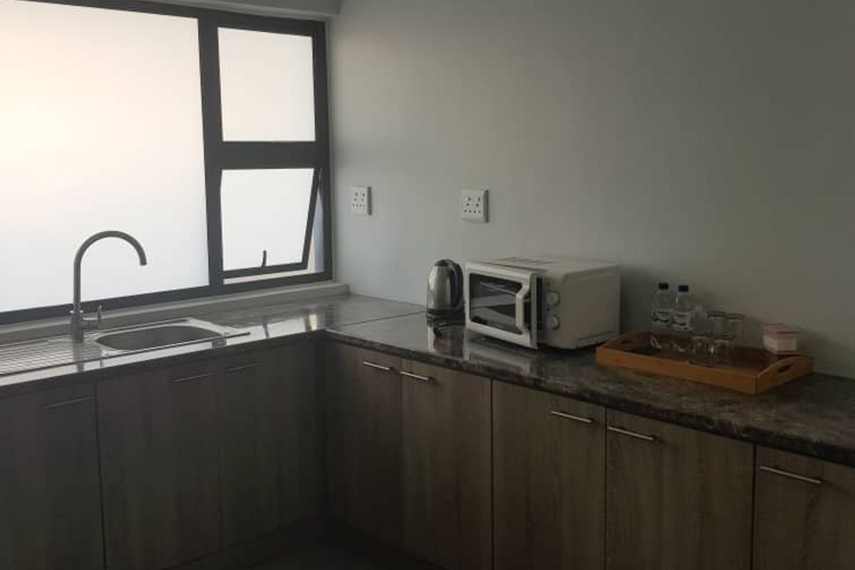 A functional kitchenette featuring wood-look cabinetry, stone-effect countertops, a stainless steel sink, and tiled flooring in a clean, neutral setting.