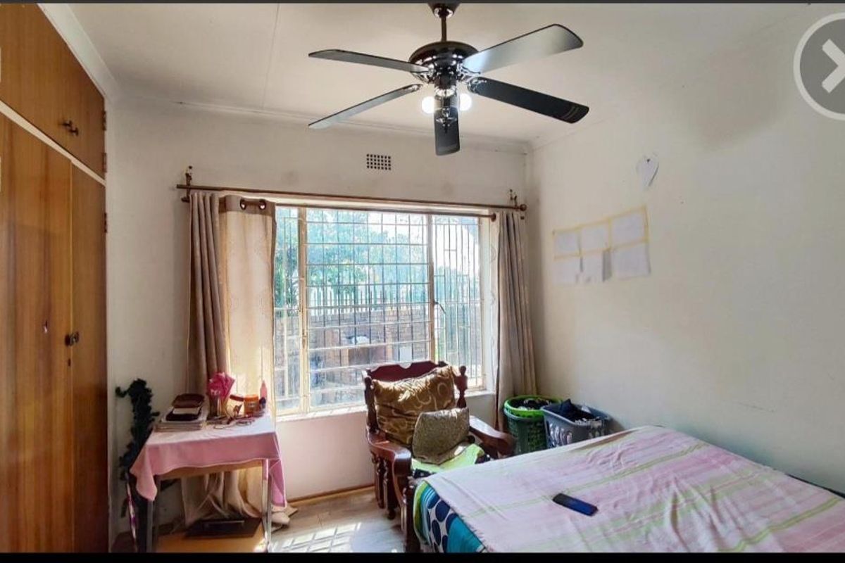 A bedroom featuring built-in wooden cupboards, a ceiling fan, and a large window with security bars.