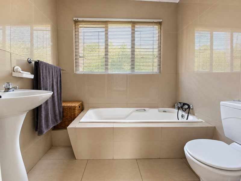 A clean, neutral-toned bathroom featuring a built-in bathtub, pedestal basin, and toilet, finished with floor-to-ceiling wall tiles and functional window blinds.