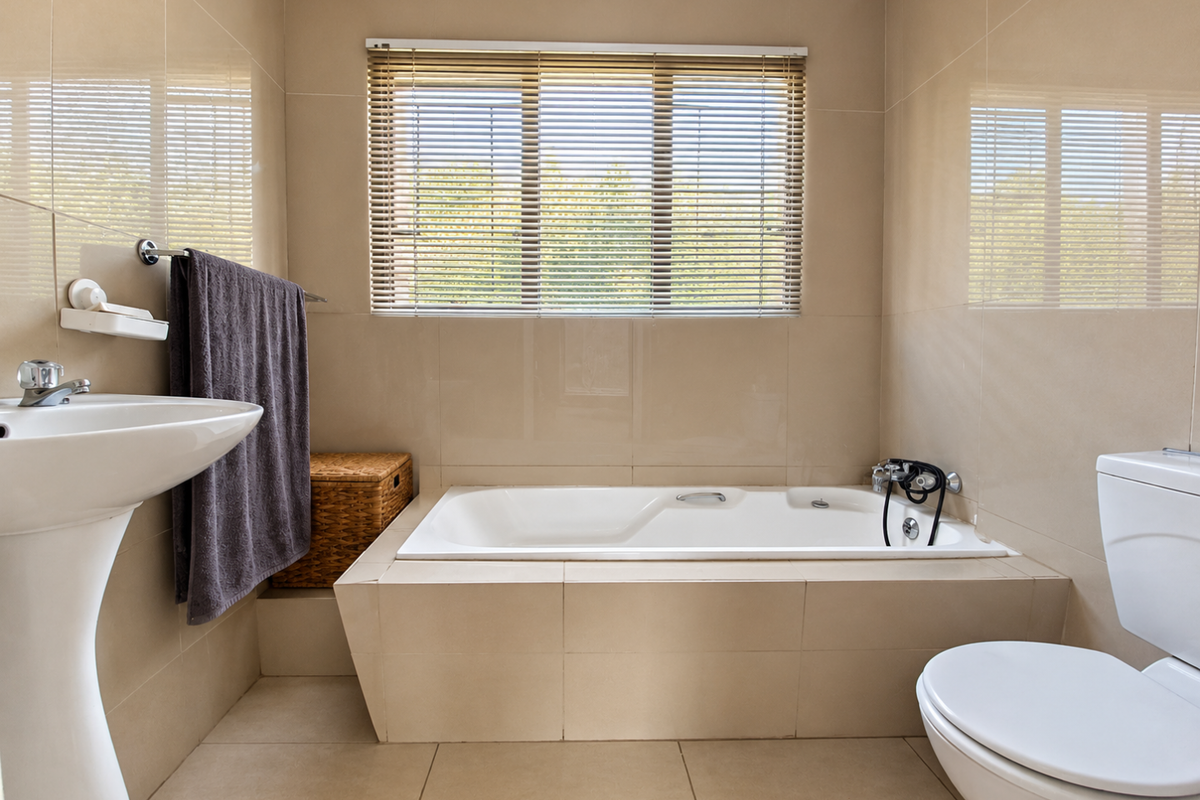 A clean, neutral-toned bathroom featuring a built-in bathtub, pedestal basin, and toilet, finished with floor-to-ceiling wall tiles and functional window blinds.
