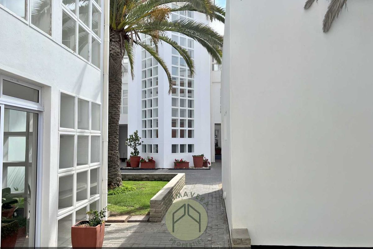 A view of the apartment complex courtyard featuring paved walkways, manicured garden beds, and modern white architectural facades with large grid-style windows.