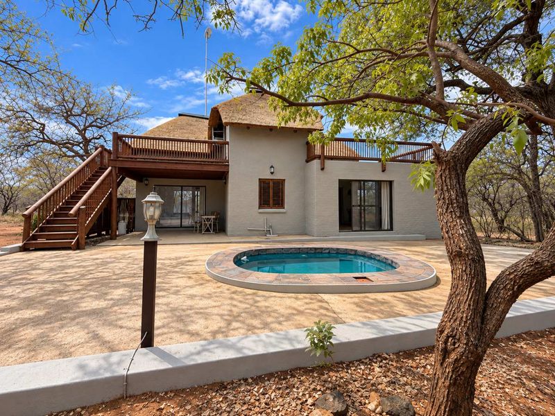 Rear view of a thatched-roof house featuring a wooden deck, external staircase, and a circular plunge pool set in a paved patio area.