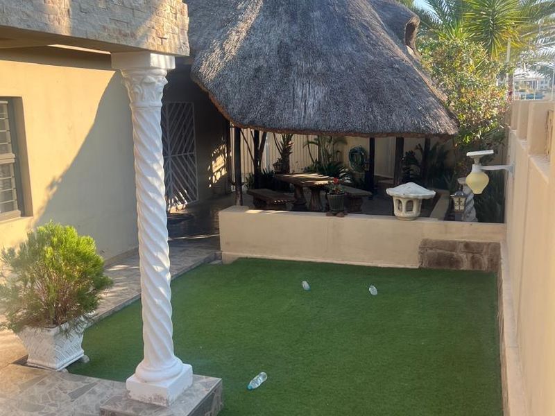 Rear garden area featuring a thatched lapa, artificial lawn, stone-clad exterior wall, and a decorative column on the tiled patio.