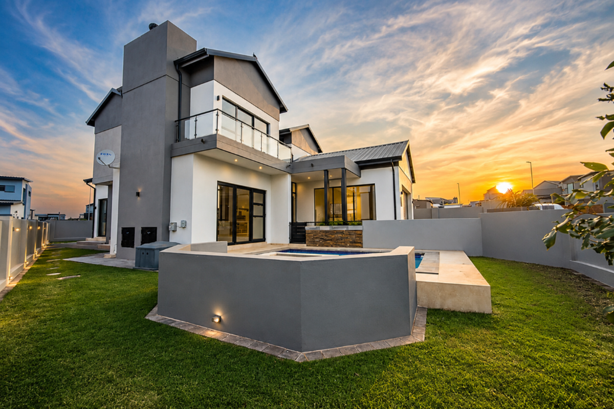 Modern double-story residence featuring a rear garden, swimming pool, stone-clad accents, and a glass-railed balcony under a sunset sky.