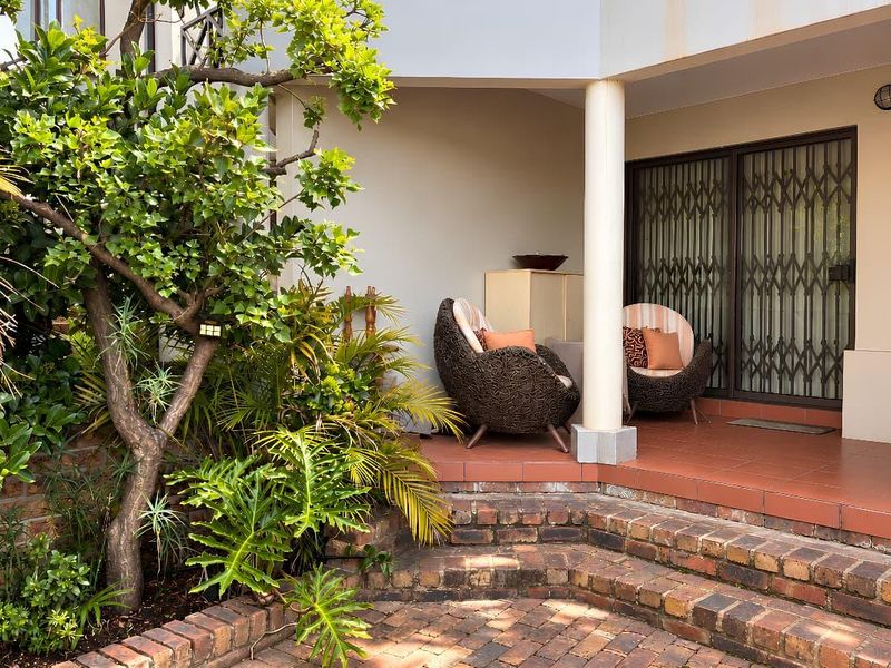 A covered patio area featuring tiled flooring, a structural column, and a secure sliding door entrance, complemented by adjacent brick paving and garden landscaping.