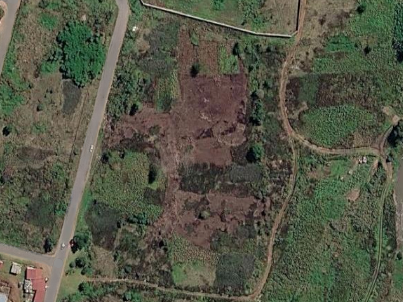 Aerial view of a vacant residential plot featuring natural vegetation and boundary fencing, situated adjacent to a paved road.