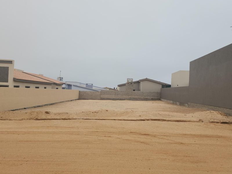 A vacant residential plot of land featuring sandy ground and enclosed by boundary walls, situated within a developed residential neighborhood.
