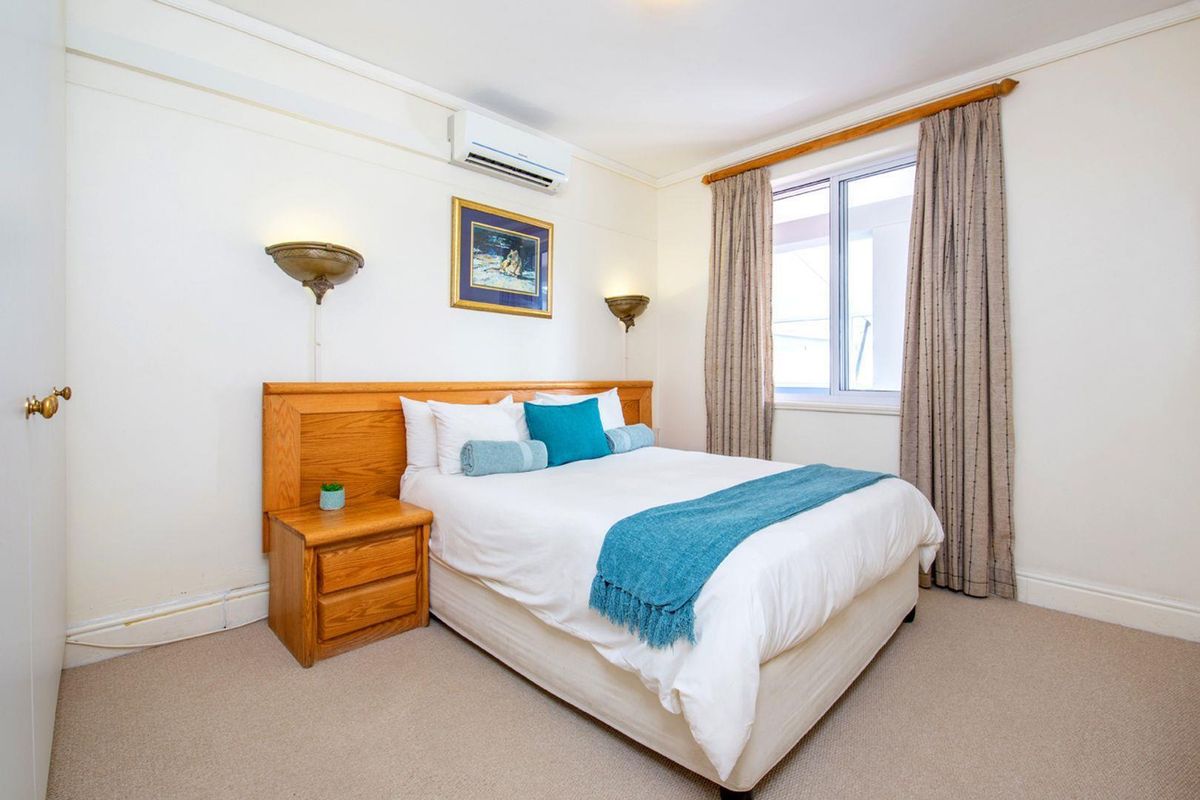 A well-maintained bedroom featuring a built-in wooden headboard, integrated bedside table, wall-mounted lighting, and a split-system air conditioning unit.