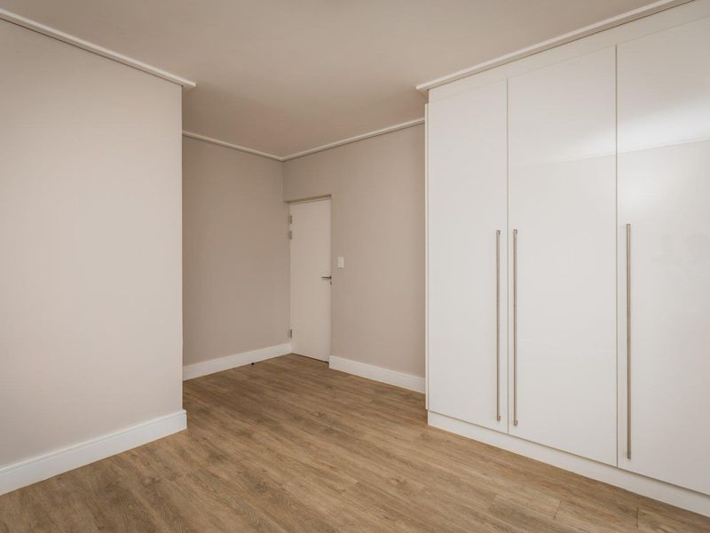 A bright, empty bedroom featuring modern laminate flooring, neutral wall tones, and sleek white built-in cupboards with long metallic handles.