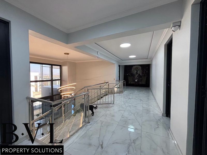 A bright, modern upper-level hallway featuring polished marble-look floor tiles, a sleek stainless steel and glass balustrade, and recessed ceiling lighting.