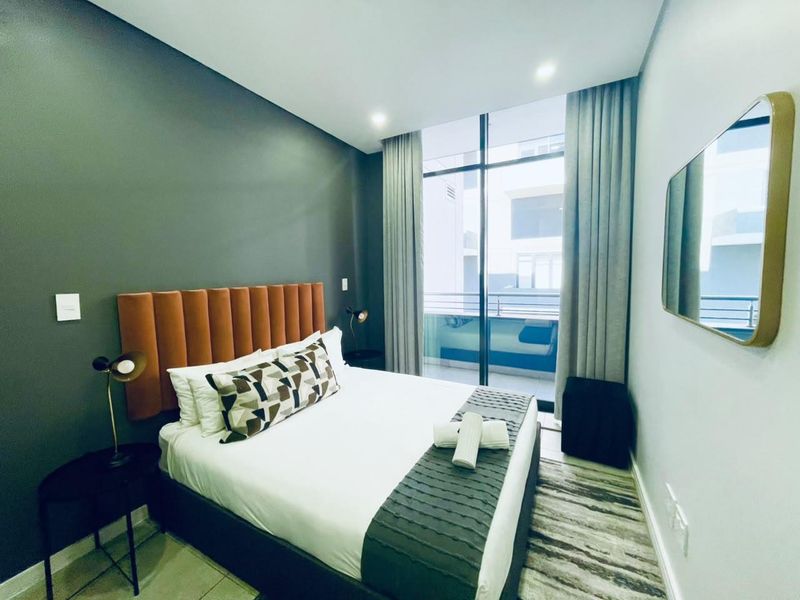 A modern bedroom featuring a dark accent wall, floor-to-ceiling windows with balcony access, and recessed lighting.