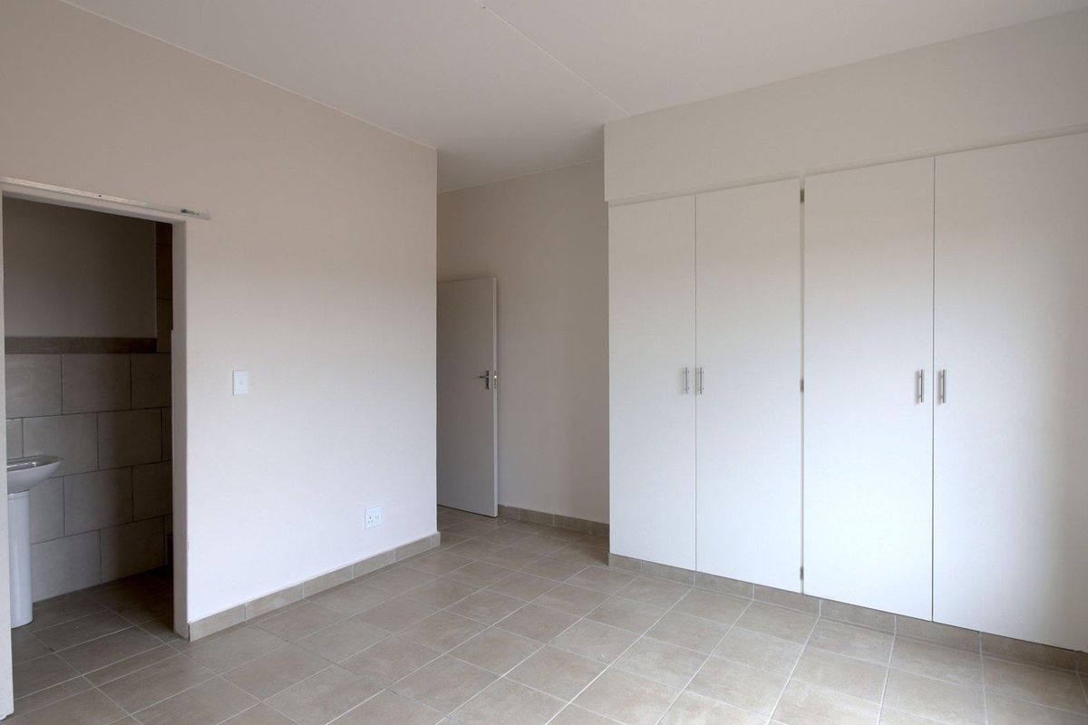 A clean, empty bedroom featuring neutral tiled flooring, ample built-in cupboard storage, and an adjoining small wash area with a pedestal basin.