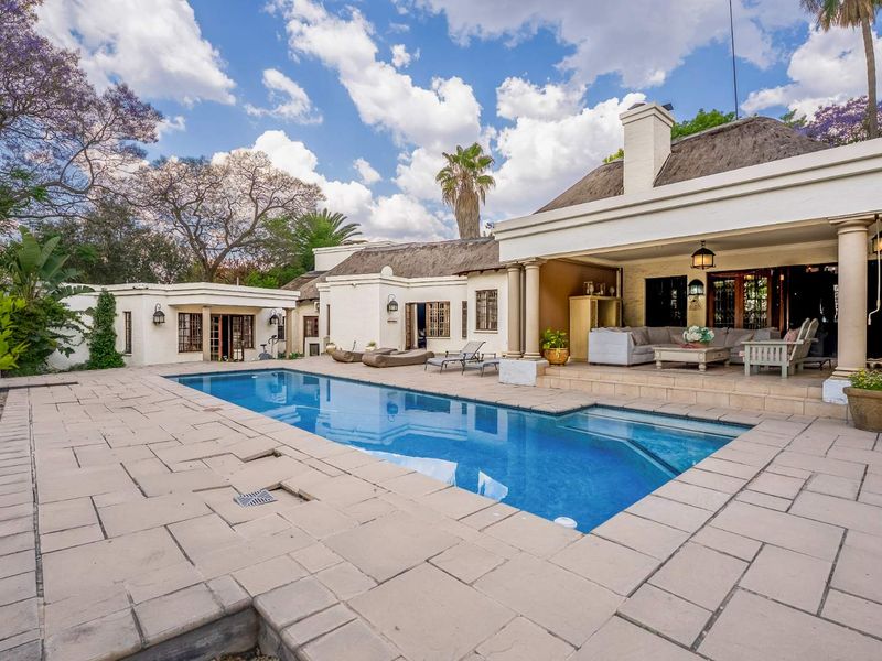A well-maintained outdoor pool area featuring a large swimming pool, stone paving, and a spacious covered patio with a thatched roof.
