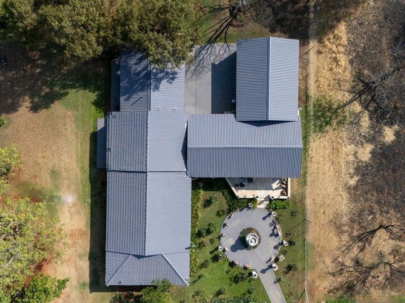 Aerial view of the farm property showcasing the main dwelling with a metal roof, paved patio area, and landscaped circular garden feature.