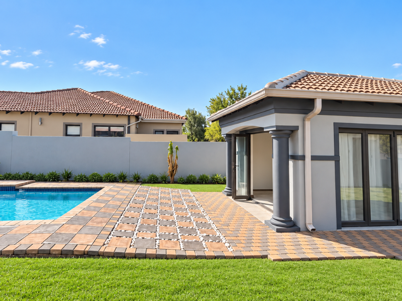 A well-maintained outdoor pool area featuring a paved patio, landscaped lawn, and a modern structure with bi-fold doors and decorative columns.