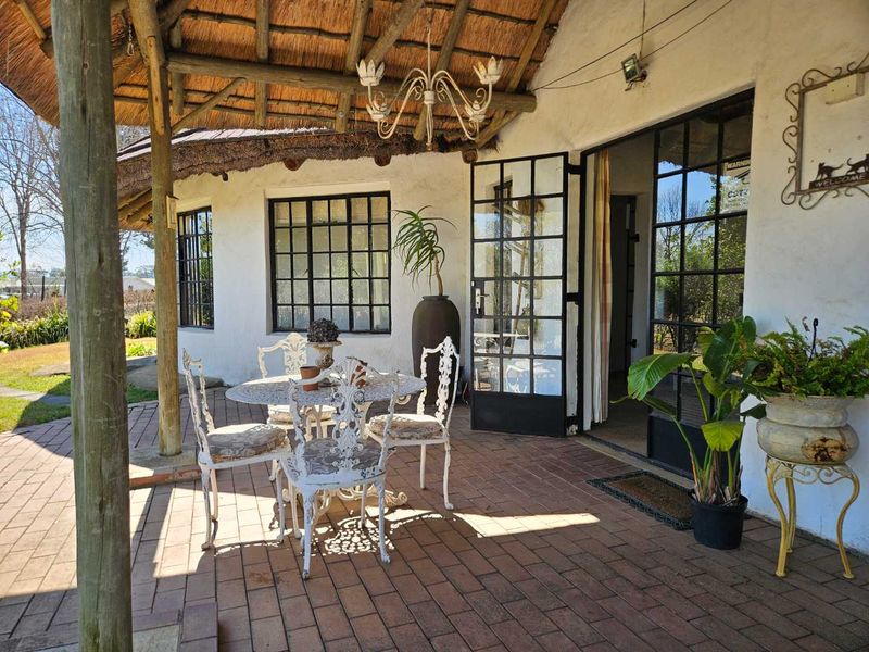 A covered outdoor patio area featuring a traditional thatched roof, brick paving, and steel-framed glass doors leading into the main dwelling.