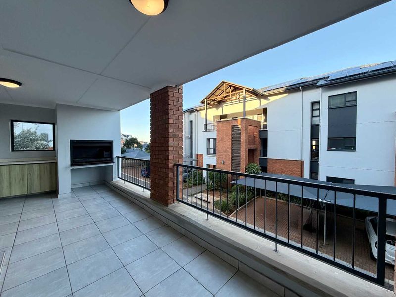 A spacious covered balcony featuring a built-in braai, tiled flooring, and a metal balustrade, overlooking the apartment complex grounds.