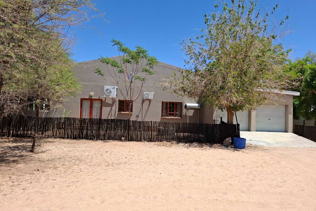Rear view of the property featuring a single-story facade, double garage, and exterior air conditioning units set within a sandy garden area.