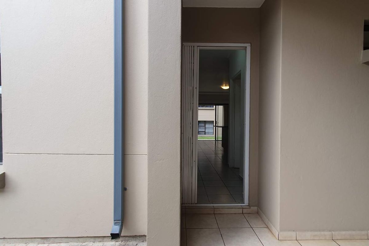A view of the rear exterior entrance featuring a security gate, tiled patio area, and a downpipe against a neutral-toned wall.