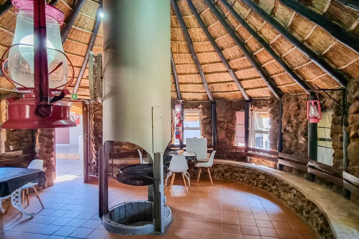 A rustic circular entertainment area featuring a central built-in fireplace, exposed timber roof structure with thatch, and integrated stone wall seating.