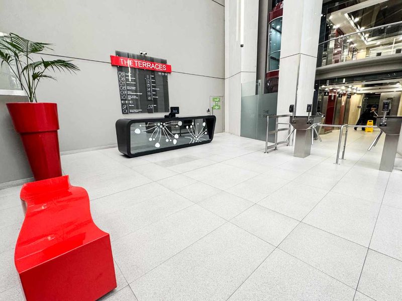 Modern office building lobby featuring a reception desk, security turnstiles, and elevator access with clean tiled flooring and professional signage.