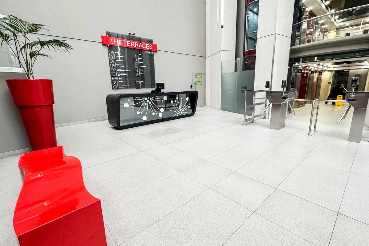 Modern office building lobby featuring a reception desk, security turnstiles, and elevator access with clean tiled flooring and professional signage.