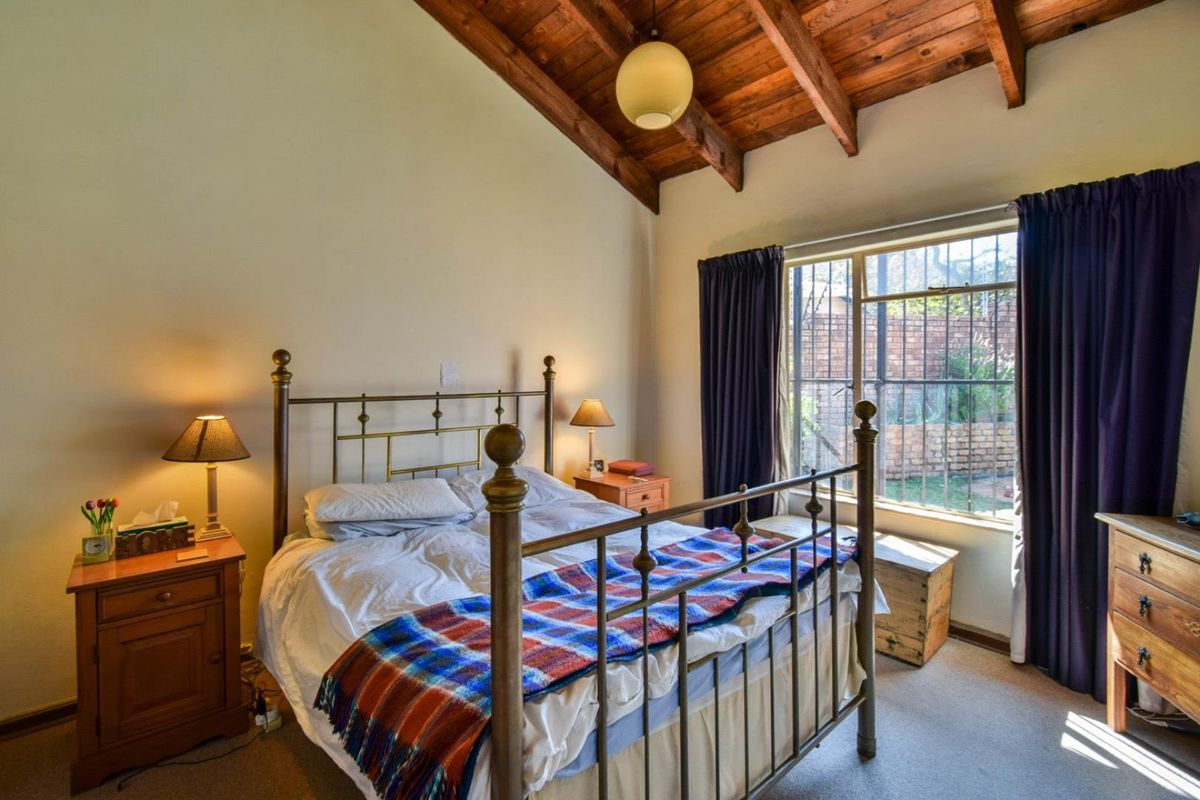 A cozy bedroom featuring a high vaulted timber-beamed ceiling, carpeted flooring, and a window with security bars overlooking the rear garden.