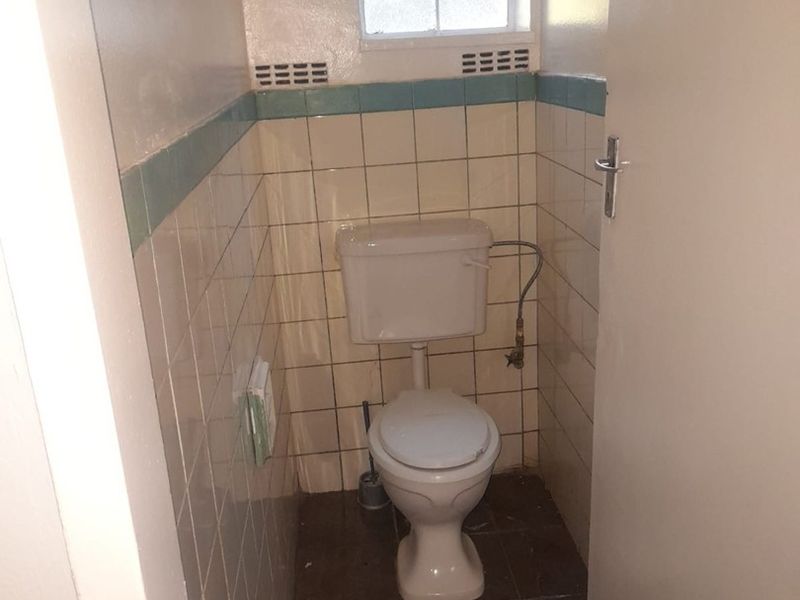 A compact, dated guest toilet featuring tiled walls and flooring, equipped with a standard toilet suite and a small window with security bars.