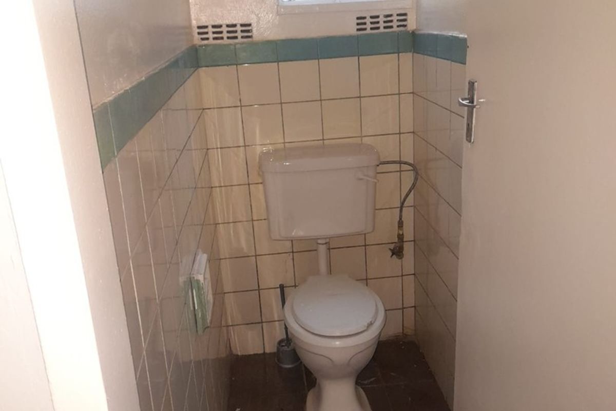 A compact, dated guest toilet featuring tiled walls and flooring, equipped with a standard toilet suite and a small window with security bars.