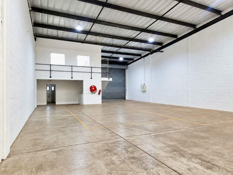 Spacious industrial warehouse unit featuring high ceilings with exposed steel trusses, concrete flooring, a roller shutter door, and a functional mezzanine level.