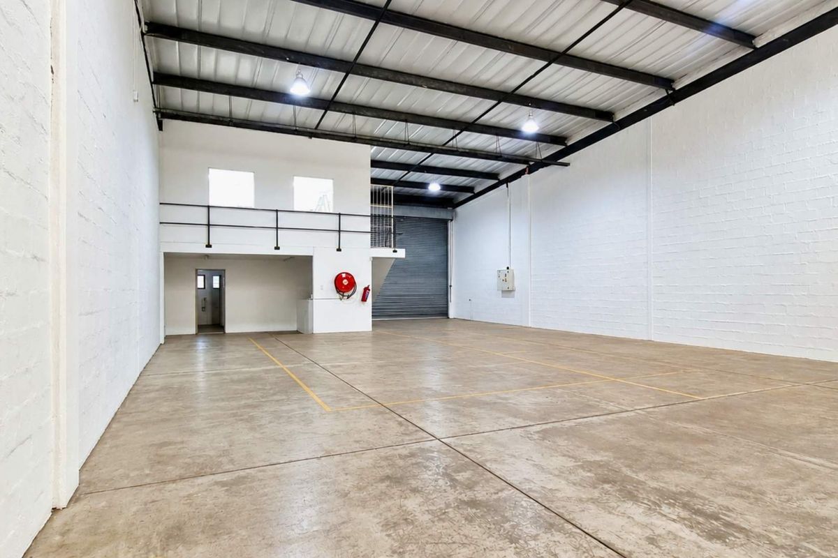 Spacious industrial warehouse unit featuring high ceilings with exposed steel trusses, concrete flooring, a roller shutter door, and a functional mezzanine level.