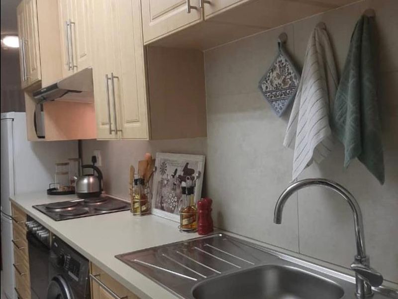 A functional kitchen featuring light wood-finish cabinetry, laminate countertops, a stainless steel double sink, and integrated appliances including an oven and electric hob.