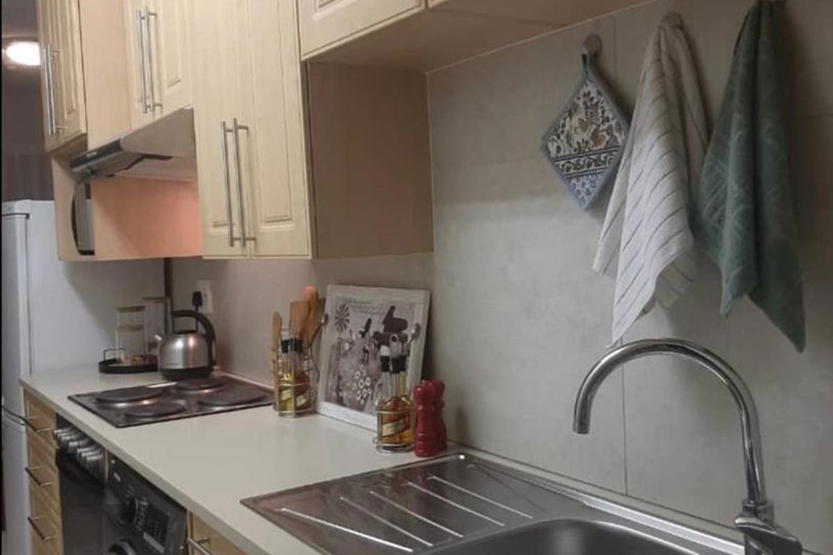A functional kitchen featuring light wood-finish cabinetry, laminate countertops, a stainless steel double sink, and integrated appliances including an oven and electric hob.