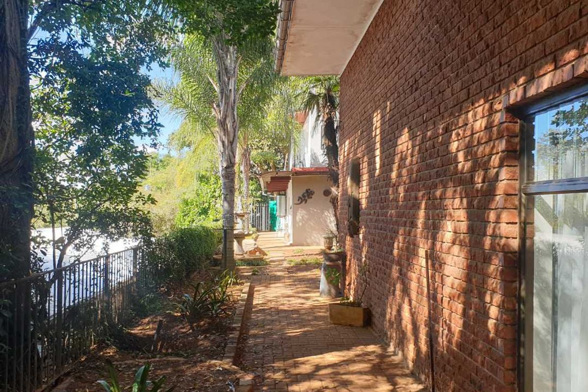 A side walkway featuring a red brick exterior wall, paved path, and metal fencing, bordered by mature trees and garden landscaping.
