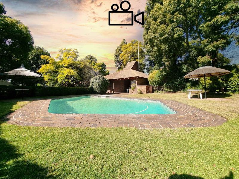 A well-maintained outdoor pool area featuring a swimming pool with brick coping, surrounded by a lush garden and a traditional thatched lapa.