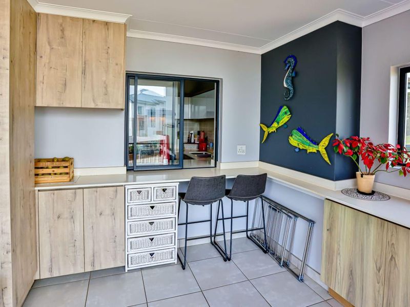 A modern kitchen area featuring light wood-finish cabinetry, laminate countertops, tiled flooring, and a convenient serving window connecting to the adjacent space.