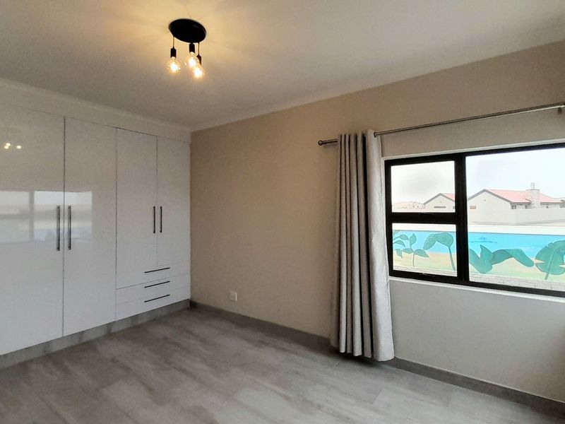 A bright, empty bedroom featuring modern white built-in cupboards, light-toned laminate flooring, and large aluminium-framed windows.