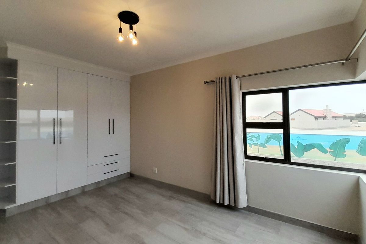 A bright, empty bedroom featuring modern white built-in cupboards, light-toned laminate flooring, and large aluminium-framed windows.