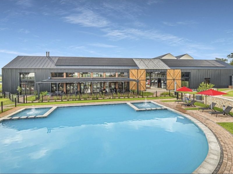 A well-maintained swimming pool area featuring water fountains, paved decking, and a modern building with solar panels and a covered patio.
