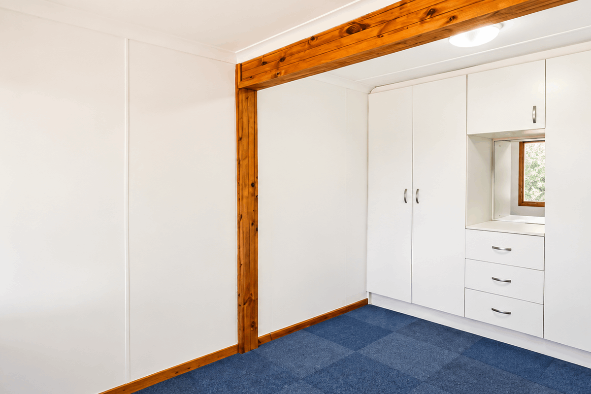 A clean, empty bedroom featuring built-in white cabinetry, a central timber ceiling beam, and blue carpet flooring.