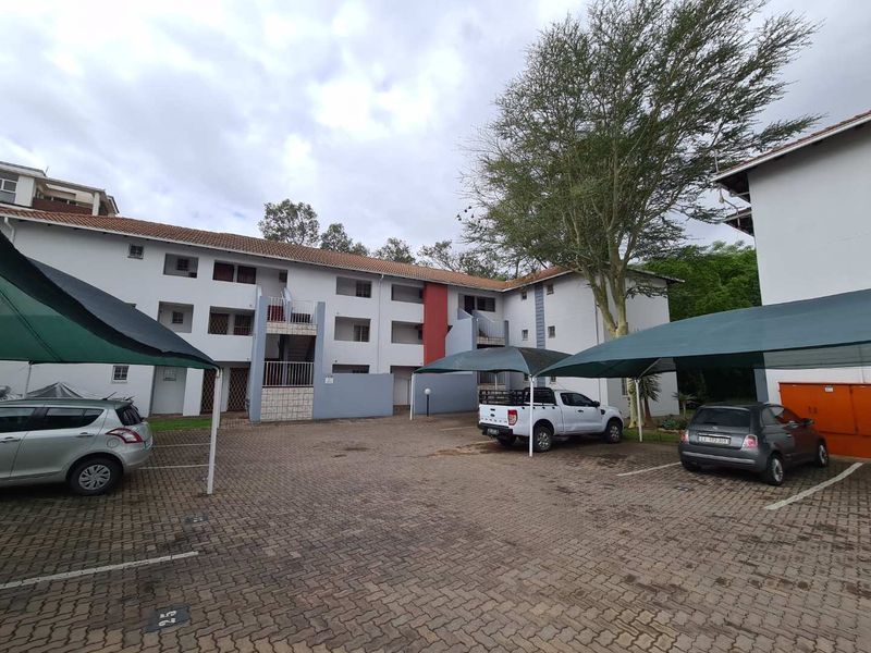 Exterior view of an apartment complex featuring a paved parking area with covered carports and multi-story residential units.