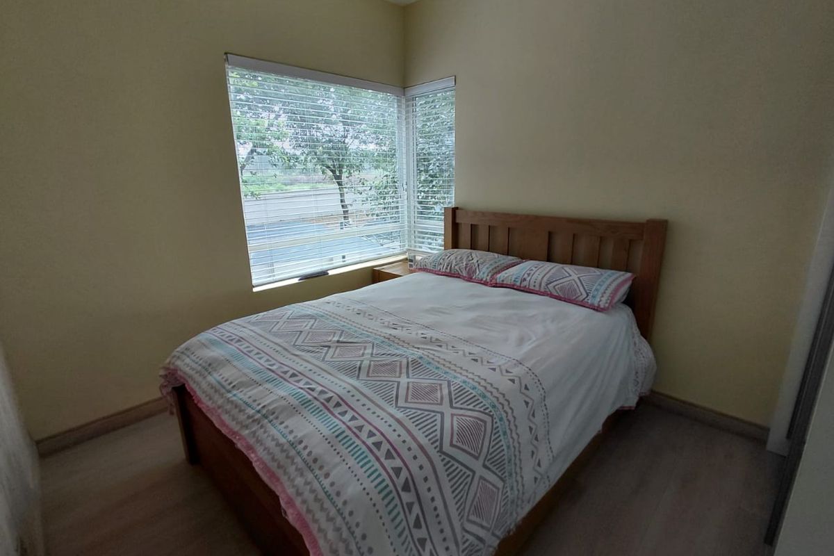 A cozy bedroom featuring light-toned laminate flooring, neutral wall paint, and a corner window arrangement that provides natural light.