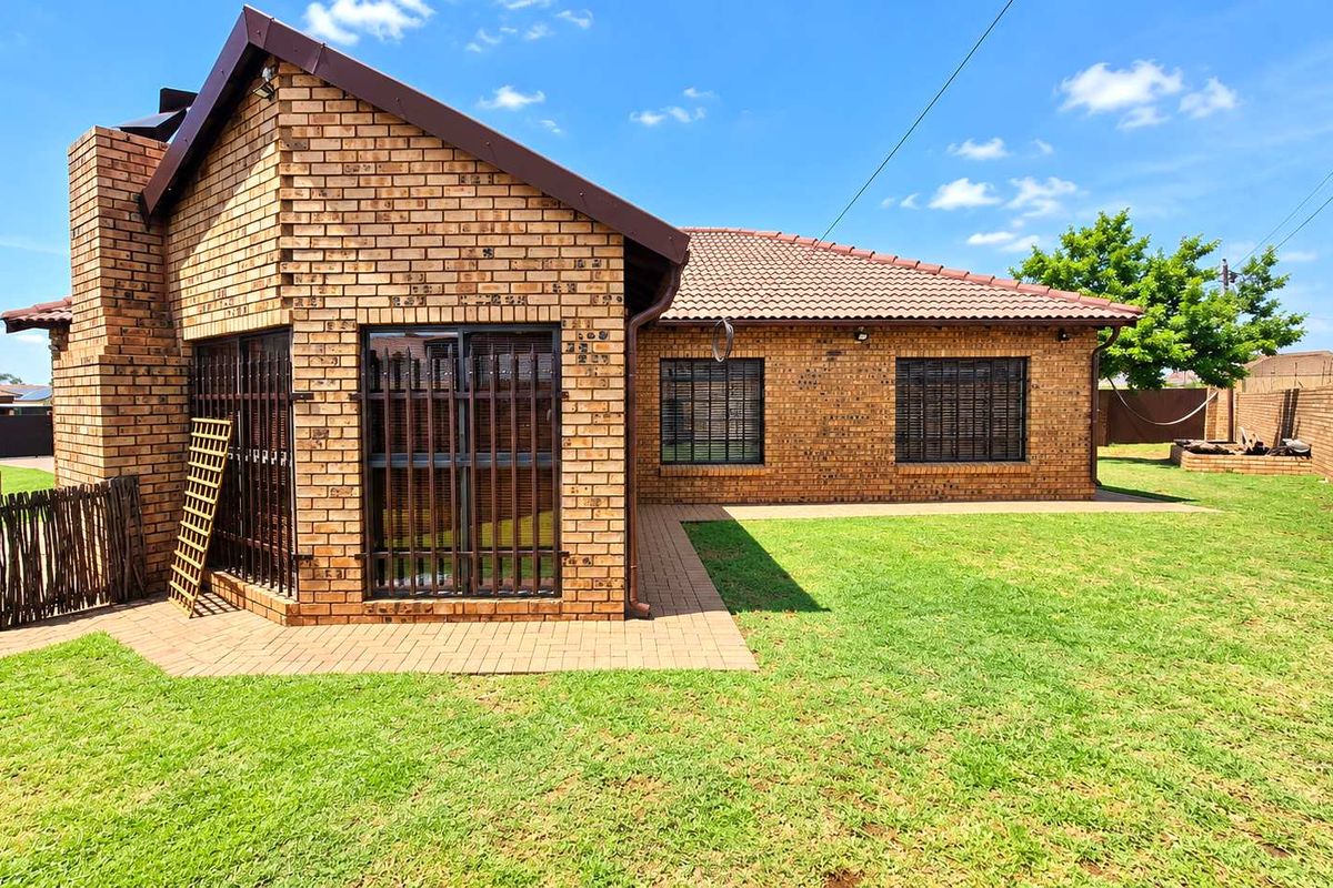 A well-maintained rear exterior view of a face brick house featuring a paved patio area, secure window bars, and a spacious private lawn.