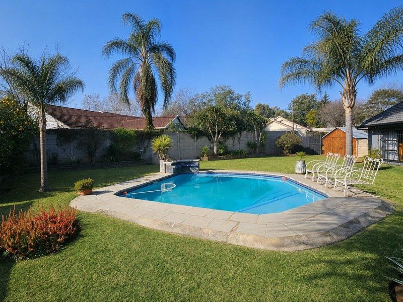A well-maintained backyard featuring a clear swimming pool with a paved surround, set within a spacious, landscaped garden with established palm trees.