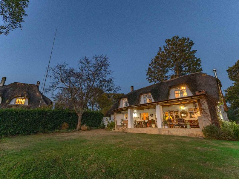 A charming thatched-roof guest house featuring a spacious covered patio overlooking a well-maintained garden at dusk.