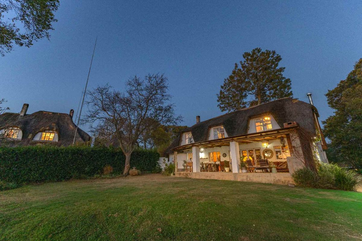 A charming thatched-roof guest house featuring a spacious covered patio overlooking a well-maintained garden at dusk.