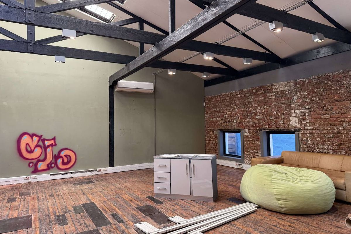 Spacious open-plan office area featuring exposed timber roof trusses, brick feature walls, skylights, and hardwood flooring, offering a unique industrial aesthetic.