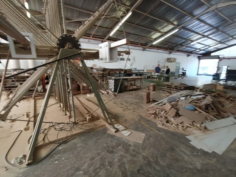 Spacious industrial warehouse area featuring exposed steel roof trusses, concrete flooring, and high-bay fluorescent lighting, currently utilized for storage and workshop activities.