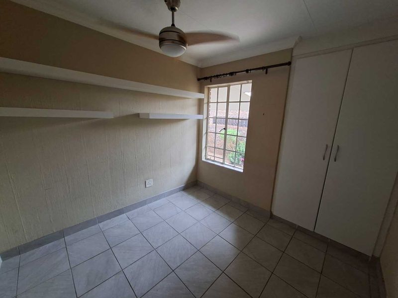 A compact bedroom featuring tiled flooring, built-in cupboards, floating wall shelves, and a ceiling fan, offering a clean and functional space.