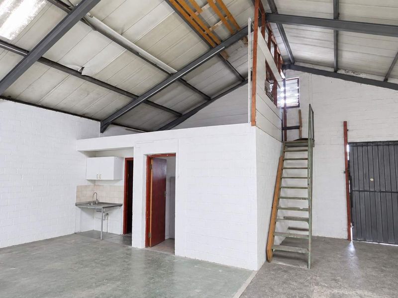 Open-plan office or industrial space featuring exposed steel trusses, concrete flooring, a small kitchenette, and a mezzanine access ladder.